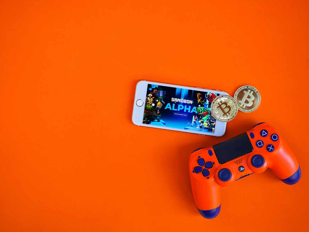 picture showing a playstation 4 controller along with a smartphone and bitcoins around it